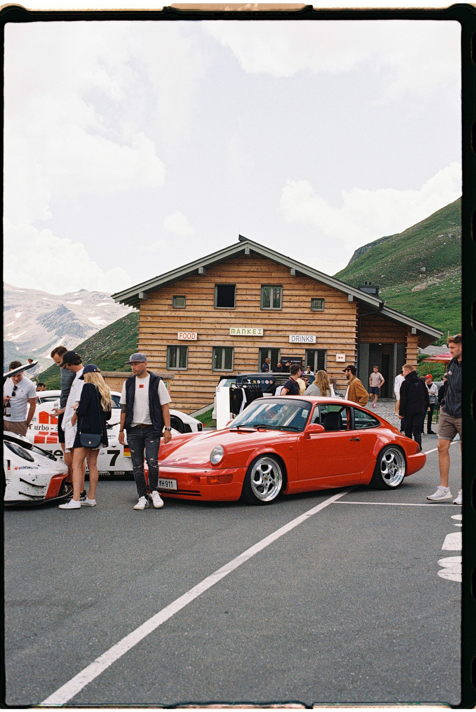 Red sports car parked in front of a wooden building with people around, mountainous landscape in the background.