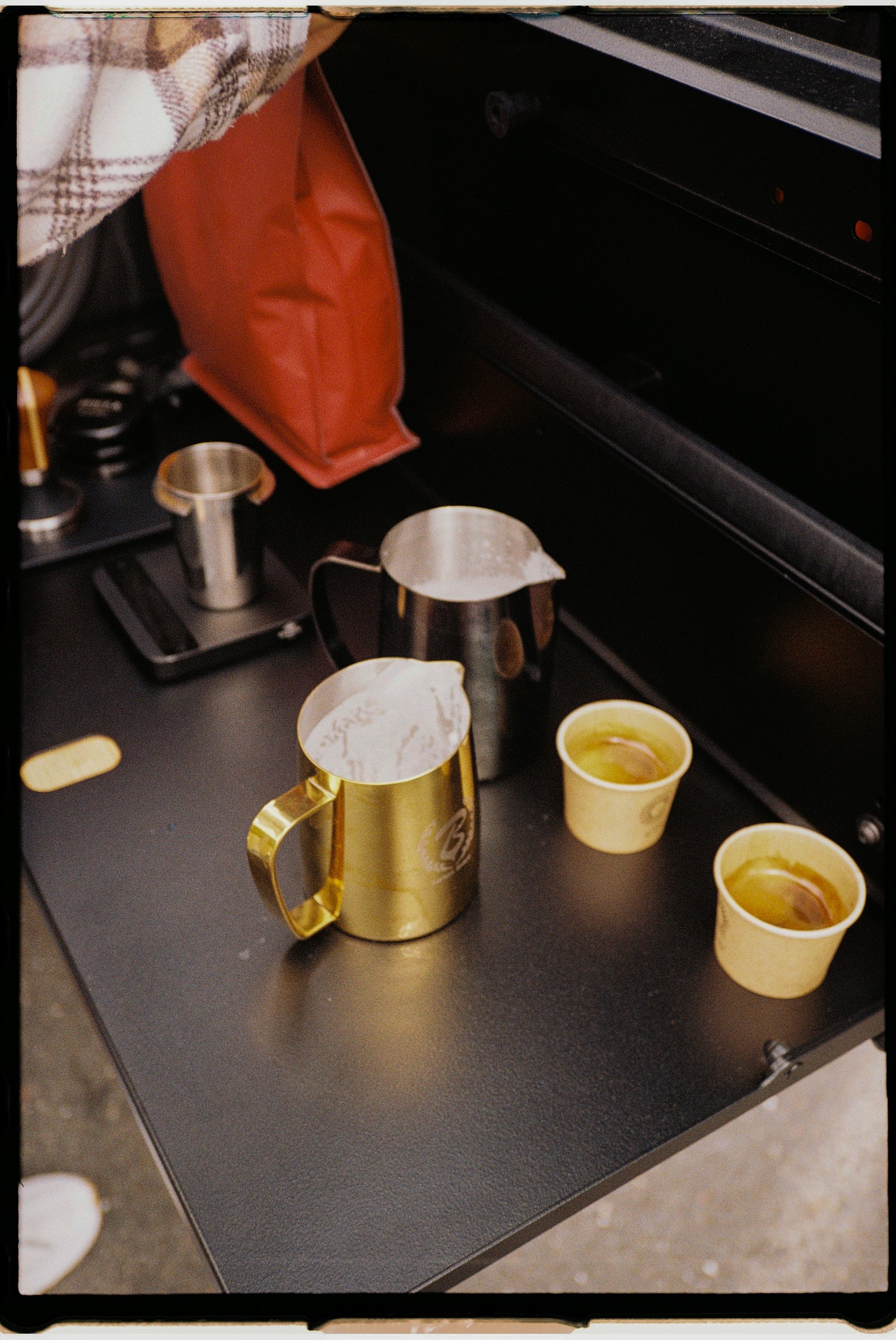 Coffee-making setup with a gold pitcher, silver frothing pitcher, and two small cups on a black surface.