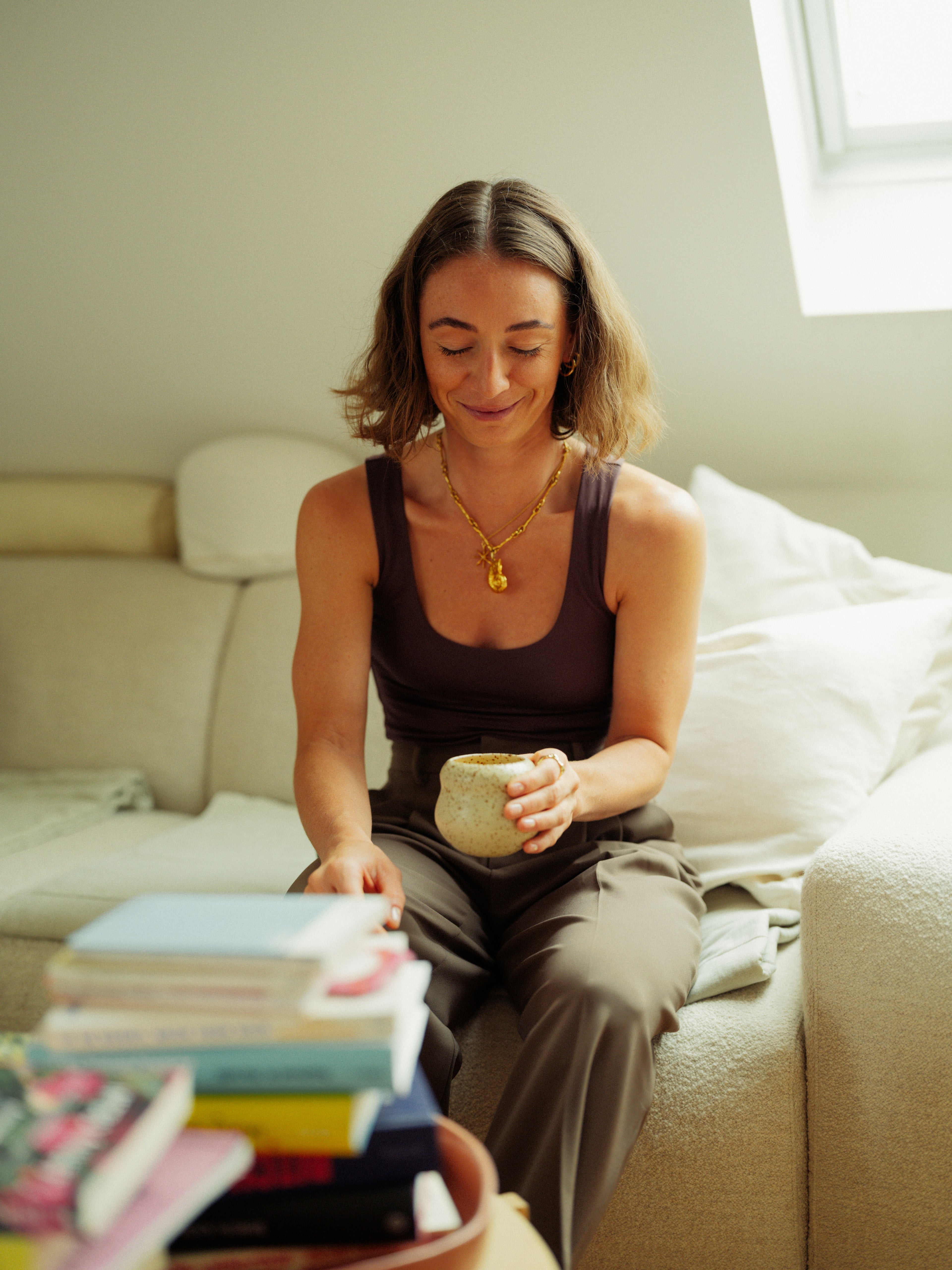 Woman sitting on a couch holding a mug with books stacked in front her on the couch table