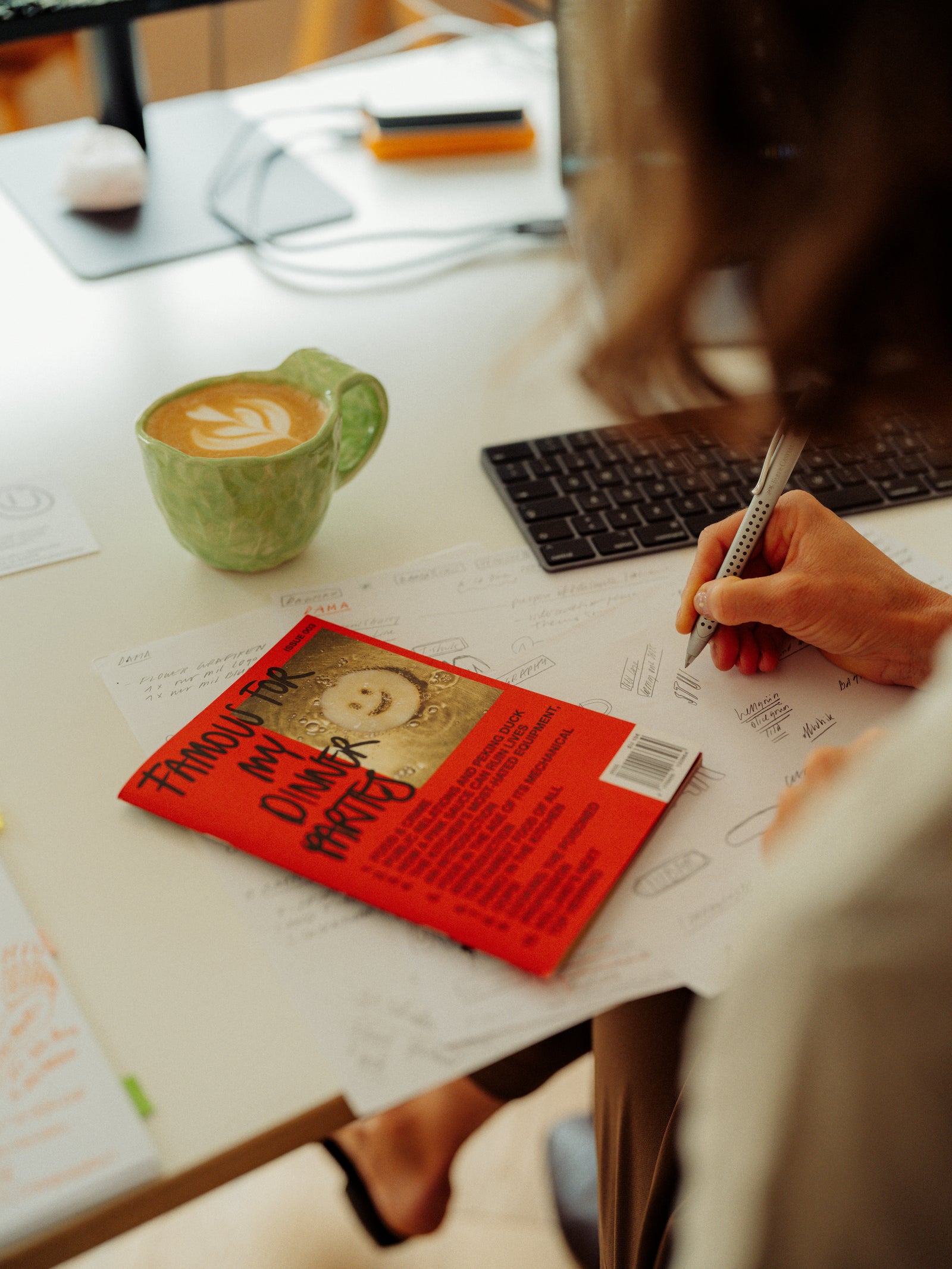 Person holding a pen over a book on a desk with a cup of coffee with latte art and computer keyboard