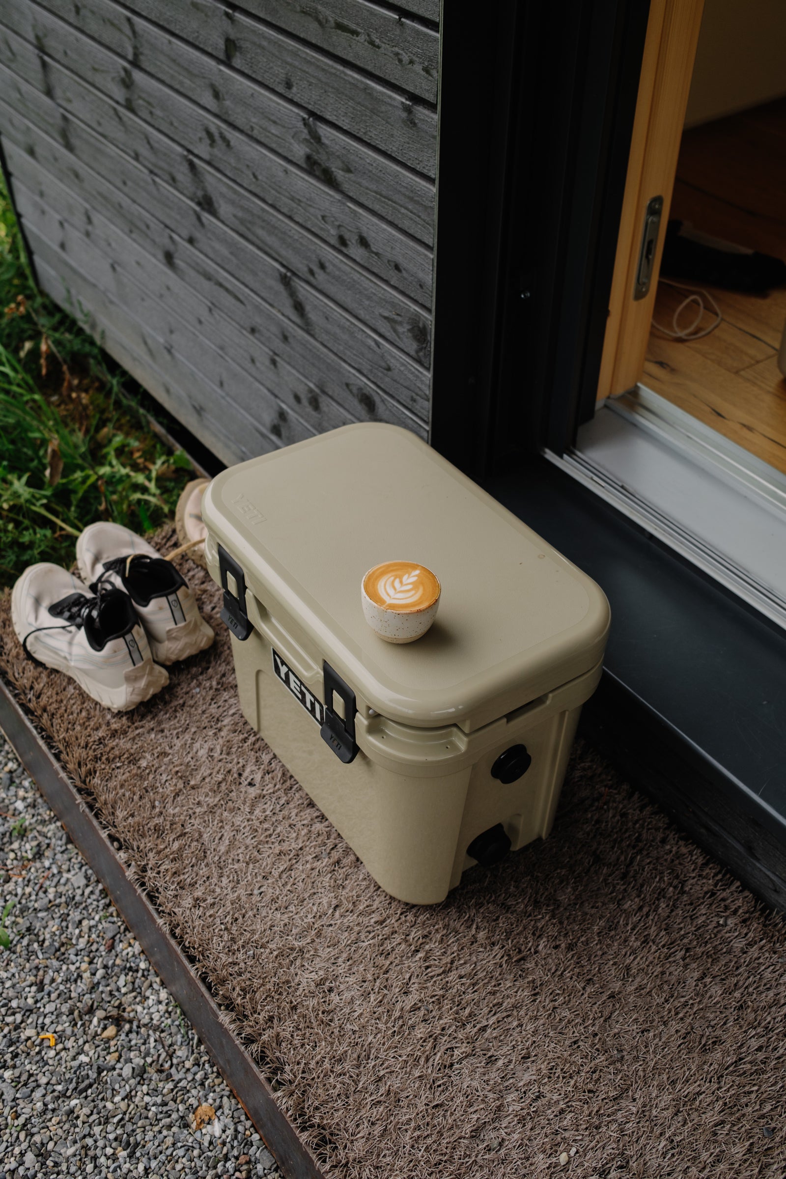 Beige Yeti cooler with a lid and  a coffee cup with latte art on top, placed against a wooden door.