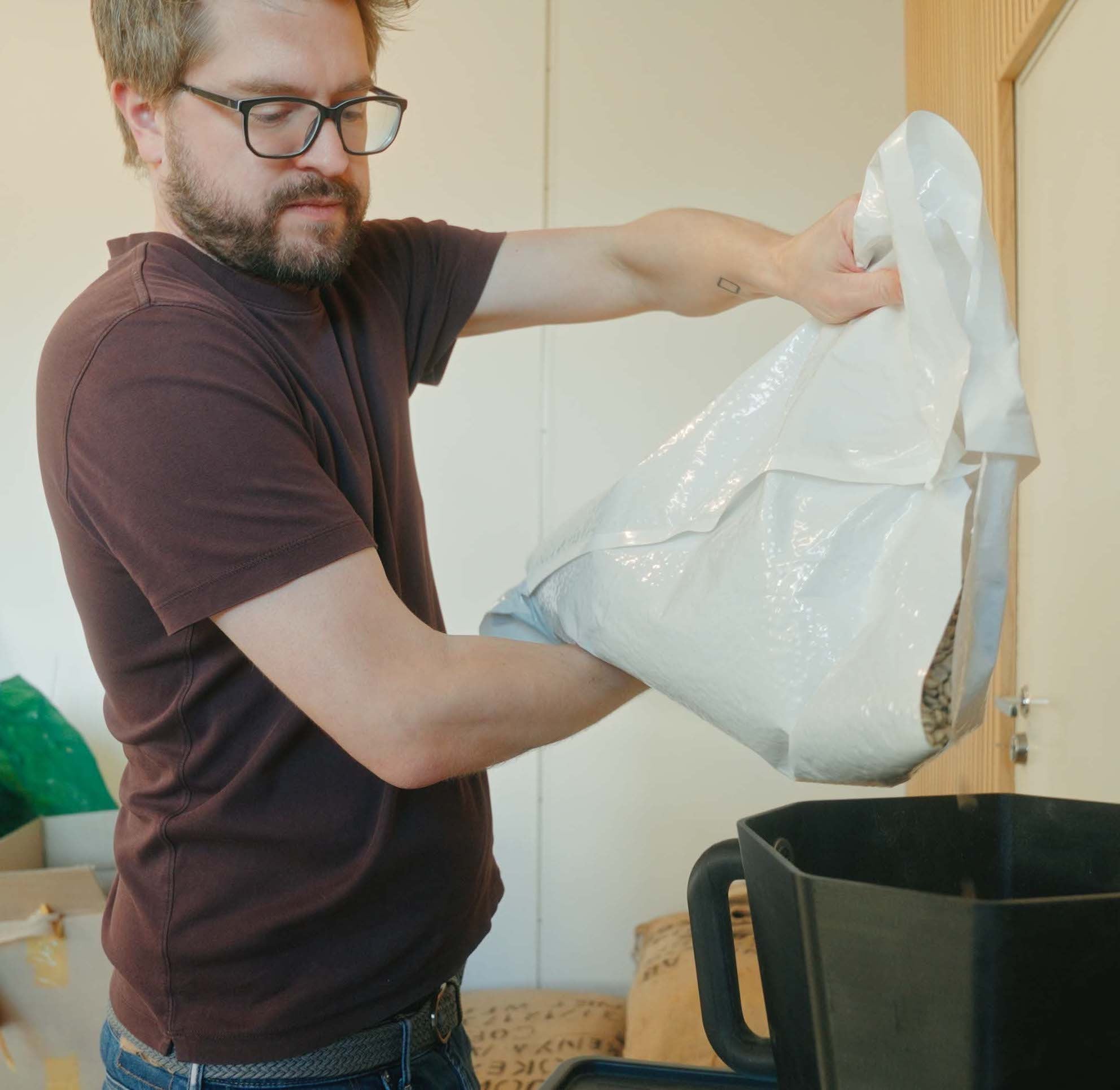 Man pouring raw coffee beans from a plastic bag into a black container indoors.
