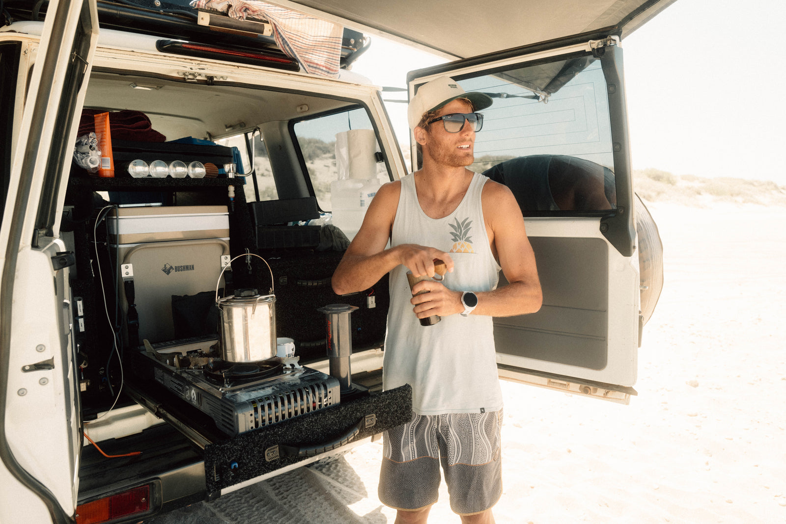 Man standing next to a van with open door, preparing Aeropress Coffee using a portable stove.