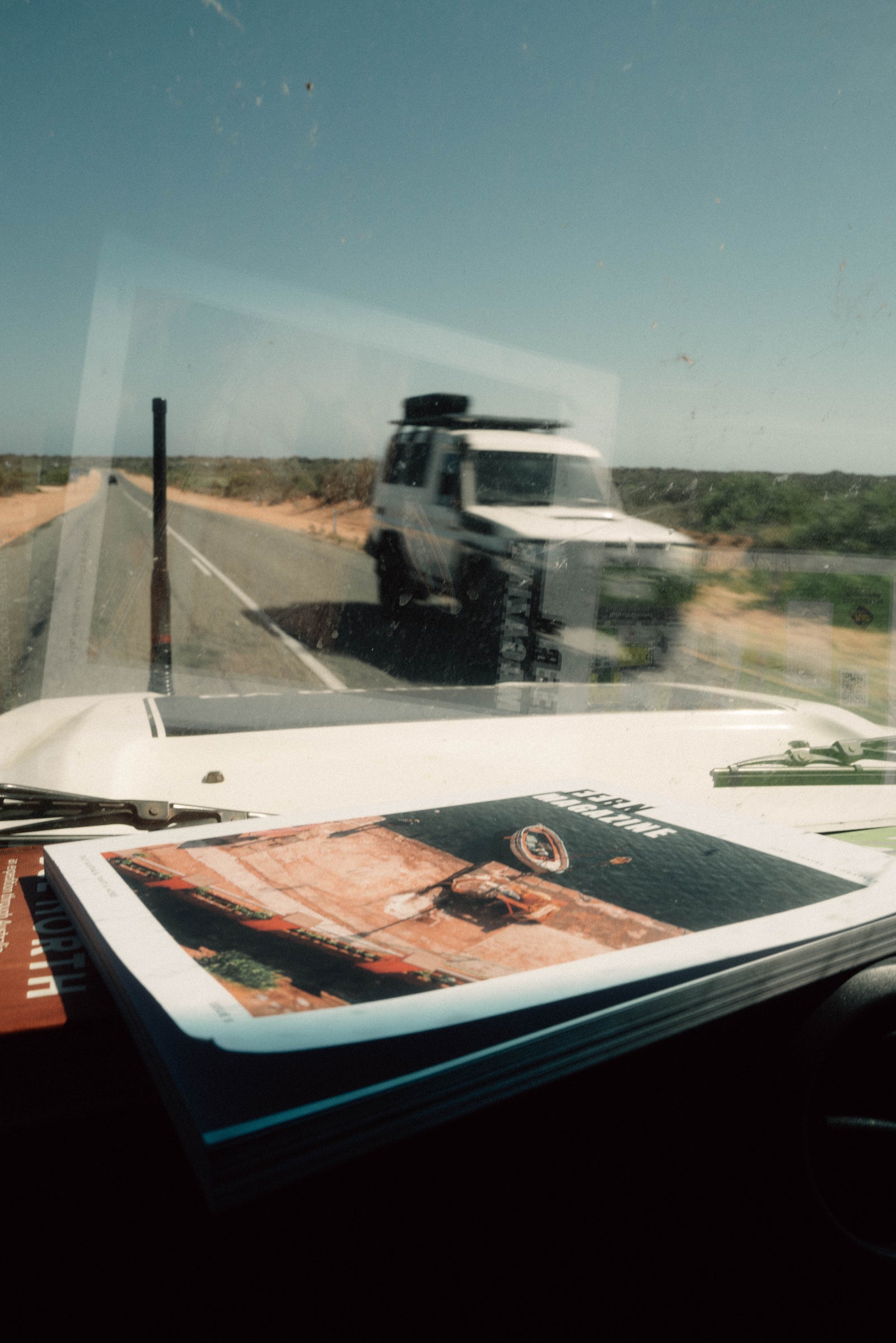 View from inside a vehicle looking out at a white SUV on a road with a clear sky with a  Fern magazine in the foreground