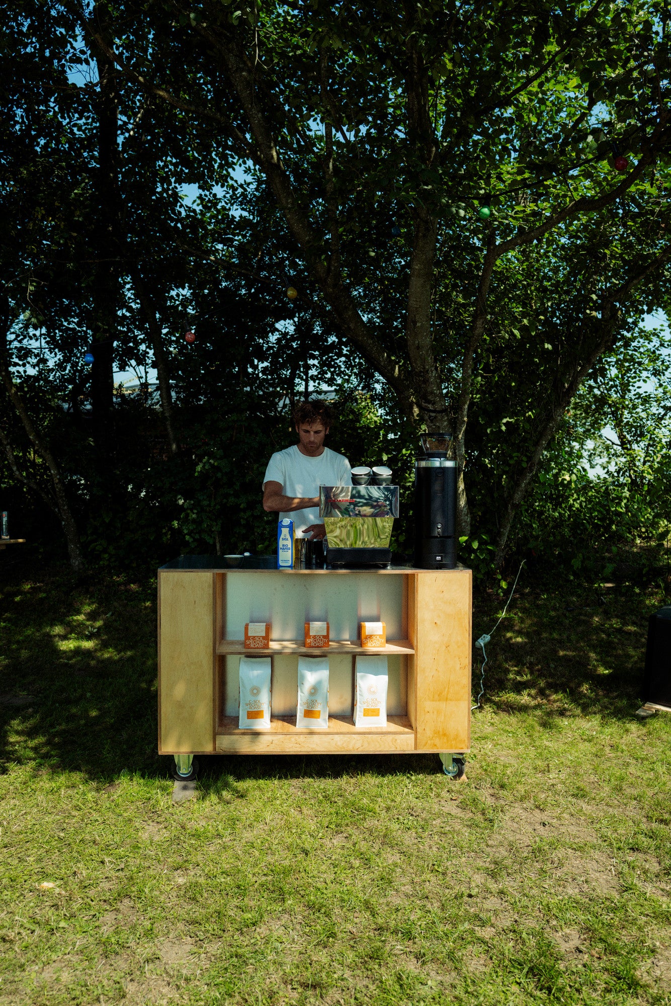 Man standing behind a wooden bar cart with drinks outdoors under trees