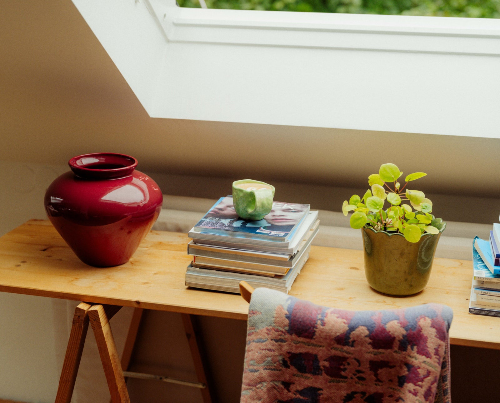 Wooden desk with a red vase, green coffee mug, and potted plant under a skylight.